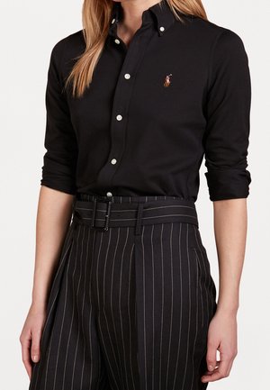 Vrouw draagt een zwart geknoopt overhemd met opgerolde mouwen en zwarte hoog getailleerde gestreepte broek met riem, torso zichtbaar.