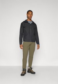Schwarze Zip-Jacke mit Kapuze, ausgestattet mit einer Brusttasche und seitlichen Reißverschlüssen, kombiniert mit olivgrünen Hosen und schwarzen Wanderschuhen.