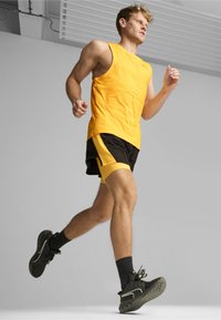 Geel sporttanktop gecombineerd met zwart en gele hardloopshorts. De hardloper draagt zwarte sneakers met witte accenten. Gladde stoftextuur.