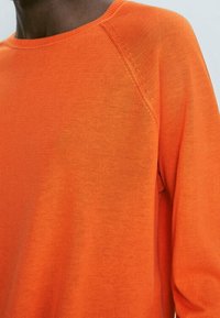 Leuchtend orangefarbener Strickpullover mit rundem Halsausschnitt, Raglanärmeln und glatter Textur, der eine lebendige Farbe mit subtilen Variationen zeigt.