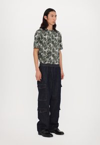 Cămașă cu model de palme verzi, cu mânecă scurtă, asociată cu pantaloni cargo de un albastru închis, având multiple buzunare și un elastic la brâu. Pantofi negri.