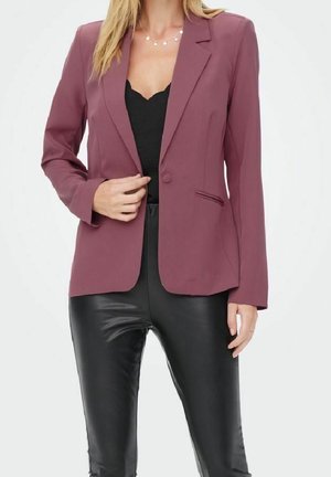 Vrouw draagt een mauve blazer over een zwart topje met een gekartelde halslijn en zwarte leren broek, houdt met één hand de voorkant van de blazer vast.