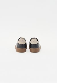 Mørkeblå lær sneakers med en tan gummisåle. Har en beige ruskinds hælflap med "CLAE" preget. Minimalistisk design, snørebåndsløs.