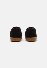 Adidași negri din suede cu talpă din cauciuc de tip gumă, având un tab de apăsare la spate și căptușeală roșie. Formă rotundă și suprafață superioară texturată.