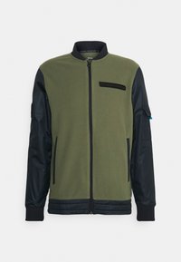 Zelená fleecová bunda s tmavě modrými nylonovými rukávy, žebrovanými manžetami a zipovým uzávěrem. Má na hrudi nášivku s logem a boční kapsy.