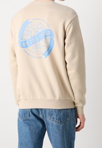 Persona che indossa un felpa beige con grafica di un globo blu e la scritta "BY LES DEUX" sulla schiena, abbinata a jeans di denim blu.