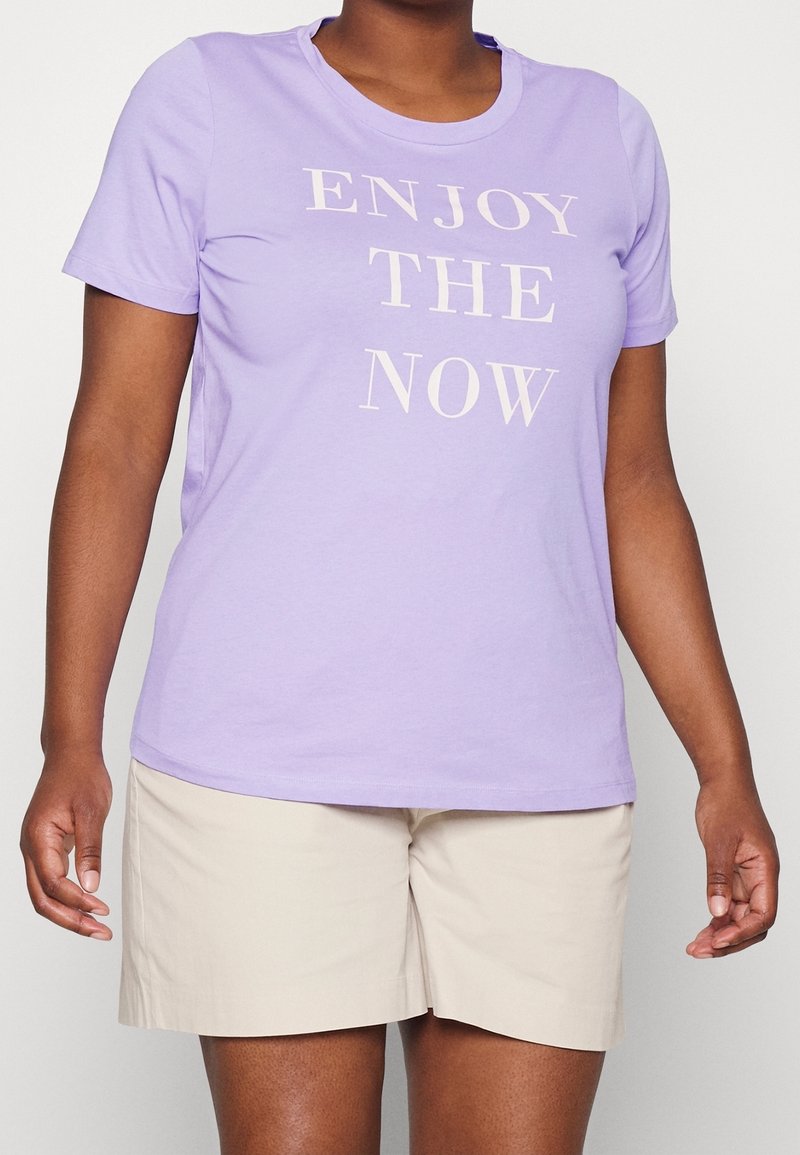 Lilla T-shirt med korte ærmer og hvid tekst, der står "NYD NUET." Fremstillet af blødt stof, stylet med beige shorts. Glat tekstur og klassisk pasform.