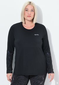 Vrouw met stijlblond haar, gekleed in een zwart sportshirt met lange mouwen en patroon op de mouwen, en bijpassende leggings, staande tegen een witte muur.