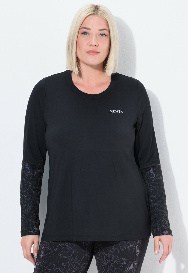 Vrouw met stijlblond haar, gekleed in een zwart sportshirt met lange mouwen en patroon op de mouwen, en bijpassende leggings, staande tegen een witte muur.