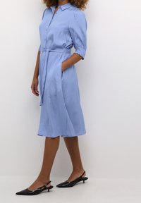 Robe chemise bleu clair à manches courtes, taille cintrée, poches latérales et jupe fluide; associée à des escarpins à bride arrière en cuir verni noir.