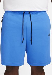 Blå idrottsshorts med svarta snoddar och sidoficka med blixtlås, bärs av en person med mörk hudfärg och vit skjorta.