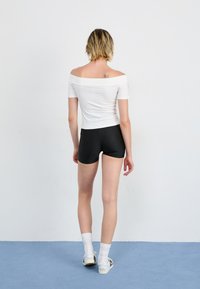Top branco de ombro a ombro e calções pretos. Modelo usa meias brancas e sapatilhas pretas e brancas. O fundo é simples com chão azul.