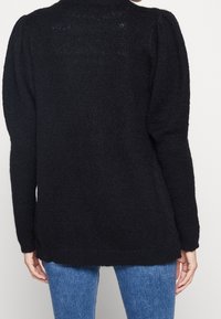 Černý pletený svetr s dlouhými rukávy, vysokým límcem a uvolněným střihem. Texturovaná tkanina a boční rozparky na spodním lemu.