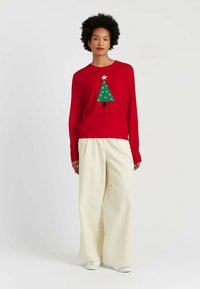 Roter Pullover mit einem grünen Weihnachtsbaumdesign, das mit Punkten und einem Stern verziert ist. Kombiniert mit weiten elfenbeinfarbenen Hosen und weißen Schuhen.