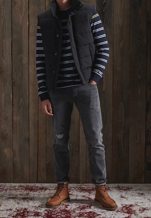 Sort puffy vest over en marineblå og stribet langærmet skjorte, kombineret med slidte grå jeans og brune støvler. Træbaggrund.