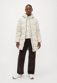 Beige pufferjakke med hætte, der har lynlås, to sidelommer og ribbet tekstur. Kombineret med mørke kordfløjl bukser og hvide sneakers.
