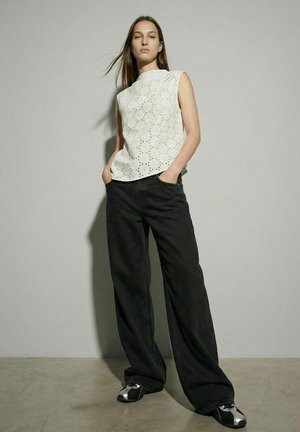 Femme debout, les mains dans les poches, portant un haut blanc sans manches en tissu ajouré, un pantalon noir ample et des baskets noir et blanc sur fond gris.