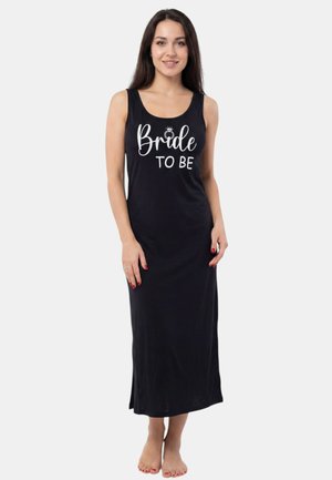 Junge Frau mit langen dunklen Haaren, die ein ärmelloses schwarzes Kleid mit dem Text "Bride to Be" trägt, steht barfuß auf hellem Hintergrund.