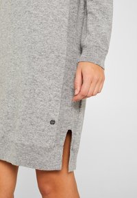 Vestito grigio a maglia con texture a costine, caratterizzato da spacchi laterali e una piccola etichetta con il marchio vicino all'orlo. Maniche lunghe e taglio morbido.