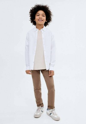 White button-up shirt over beige top, paired with brown pants and gray sneakers. Background is plain, focusing on the outfit and colors.