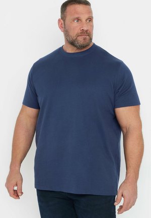 Skæggede mand iført en blå ribstrikket kortærmet t-shirt og mørke bukser, kiggende til venstre mod en ensfarvet hvid baggrund.