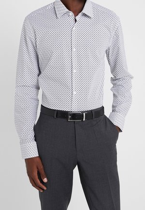 Mężczyzna w białej koszuli na guziki z powtarzającym się ukośnym wzorem tekstu, ciemnoszarych spodniach i czarnym pasku z srebrną klamrą.