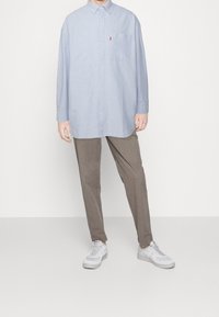 Henkilö, jolla on päällään ylisuuri vaaleansininen nappipaita, ruskeat kapenevat housut ja valkoiset urheilukengät, seisoo tasaisella valkoisella taustalla.