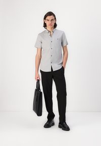 Grijze button-up shirt met korte mouwen en een borstzak, gecombineerd met zwarte broek en zwarte schoenen, terwijl hij een zwarte leren tote bag vasthoudt.