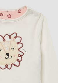 Langsleeve crèmekleurig shirt met een gestikt leeuwengezicht grafiek met een bruine rand. Drukknoopsluiting bij de schouder en een roze bloemenpatroon aan de binnenkant van de nek.