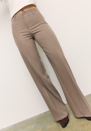 Beige, hoch taillierte, weit geschnittene Hose, getragen mit braunen spitzen Schuhen vor einem weißen Wandhintergrund.