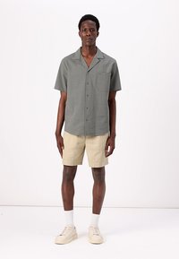 Camisa de manga corta con botones en gris texturizado, con un bolsillo de parche, combinada con pantalones cortos de lino beige y calcetines y zapatillas blancas.