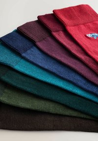 Asto adītās šalles sakārtotas kaudzē, parādot dažādas krāsas, tostarp sarkana, bordo, zila, zilgani zaļa, zaļa un brūna.