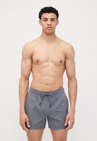 Shorts de bain gris avec un cordon de serrage à la taille, en tissu léger et à coupe droite, portés par un modèle torse nu sur un fond neutre.