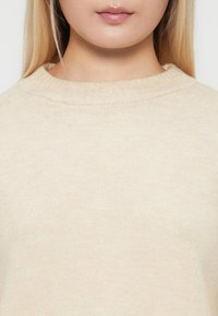 Gros plan sur une personne portant un pull beige à col ras du cou côtelé, montrant les lèvres, le menton, le cou et le haut de la poitrine avec de longs cheveux blonds visibles.