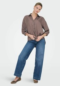 Brun skjorte med knapper og oprullede ærmer, parret med blå vide bukser og brune flade sko. Outfittet har et afslappet, uformelt design.