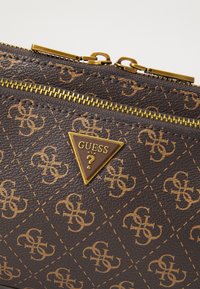Pruun tekstureeritud sünteetiline kotike kuldsete detailidega, millel on kolmnurkne Guess logo ja mustri detailid omavahel ühendatud G-dega.