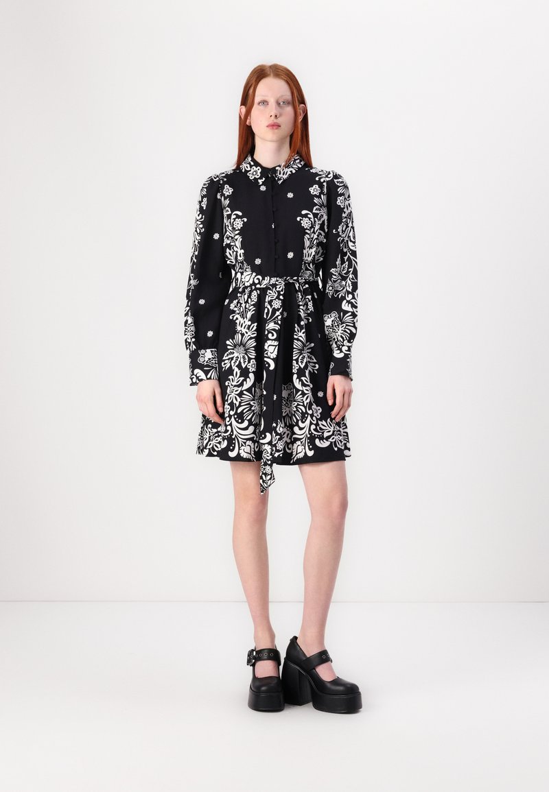 Μαύρο φόρεμα με λευκό λουλουδάτο σχέδιο, μακριά μανίκια, κουμπωτό μπροστινό μέρος και ζώνη από ύφασμα. Συνδυασμένο με χοντρές μαύρες πλατφόρμες.