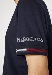 Tmavě modré tričko s krátkým rukávem z měkké bavlny. Na rukávu je bílý nápis 'HILFIGER '85' a proužky v červené, bílé a černé barvě.