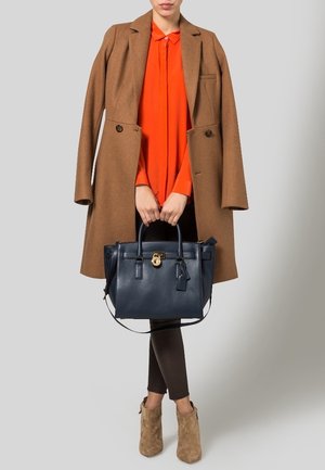 Cappotto di lana marrone, camicia arancione con bottoni, borsa in pelle blu navy con dettagli dorati, leggings marroni e stivaletti beige.