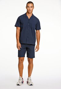 Marineblauw shirt met korte mouwen en twee voorzakken, gecombineerd met bijpassende shorts. Gemaakt met witte en marineblauwe sneakers, met een textiele stof.