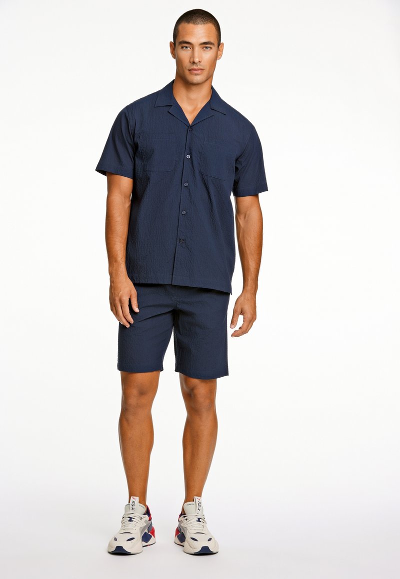 Marineblauw shirt met korte mouwen en twee voorzakken, gecombineerd met bijpassende shorts. Gemaakt met witte en marineblauwe sneakers, met een textiele stof.