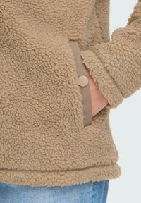 Veste en polaire beige texturée avec une main dans la poche, dotée d'une fermeture à boutons-pression et d'un jean en denim bleu clair visible en bas.
