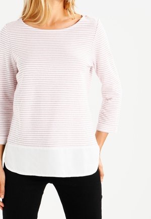 Frau trägt ein hellrosa und weiß gestreiftes Langarmshirt mit weißem, scheinbar geschichtetem Saum, kombiniert mit schwarzen Hosen.