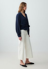 Blusa avvolgente blu navy con maniche lunghe e leggermente a sbuffo; pantaloni bianchi a gamba larga; scarpe nere senza lacci; tessuto liscio; design minimalista.