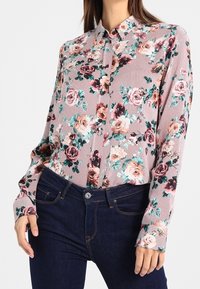 Blusa con motivo floreale su sfondo a righe, nelle tonalità di rosa, verde e nero. Design con maniche lunghe e colletto abbottonato.