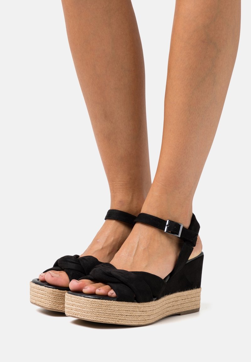 Sandales compensées noires avec une texture tressée, dessus en tissu, bride de cheville, et une semelle intercalaire enveloppée de jute. Semelle plate avec un détail minimaliste en métal.