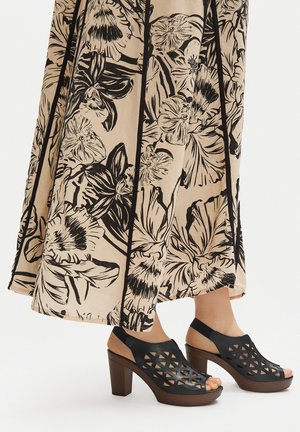 Beżowa spódnica z dużym czarnym kwiatowym wzorem, zestawiona z czarnymi, otwartymi na palce sandałami na obcasie z geometrycznymi wycięciami.
