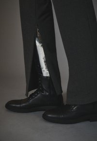 Pantalones negros con una abertura lateral que revelan una pierna prostética metálica. Combinados con botas negras de cordones que tienen una textura suave y suelas de goma.
