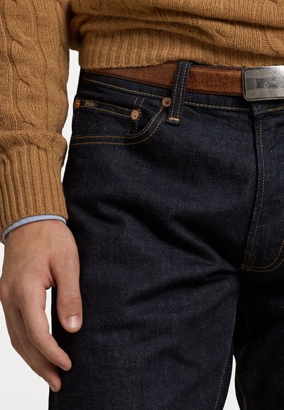 Mørkeblå denimjeans med gyldne sømmer, metallhardware og et brun lærbelte. Ribbete beige genser synlig over.