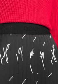 Primer plano de una blusa de punto roja metida en una falda plisada negra con texto blanco y una cinturilla negra de la marca 'HUGO'.